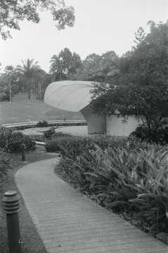 A curving pathway leads through a landscaped garden area filled with dense foliage and plants. The path approaches a modern, uniquely shaped building with a ribbed roof. Tall trees and a well-manicured lawn are visible in the background, evoking a serene and natural environment.