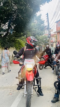A street scene featuring several motorcyclists in protective gear and helmets. The central figure is on a red bike with a visible headlight. The background includes trees, electrical wires, and residential buildings, creating an urban setting. A pedestrian walks on the roadside, moving away from the camera.