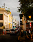 A busy urban street lined with colorful colonial buildings on both sides. The buildings showcase intricate architectural details and hues of yellow, blue, and white. Cars are lined up along the cobblestone street, and people are walking on the sidewalks. A sign for traffic, 'Transito' and 'PARE', is visible along with orange traffic cones. Overhead, there are lamps and electrical wires, and trees add greenery to the scene.