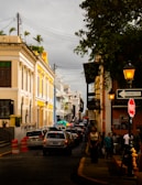 A busy urban street lined with colorful colonial buildings on both sides. The buildings showcase intricate architectural details and hues of yellow, blue, and white. Cars are lined up along the cobblestone street, and people are walking on the sidewalks. A sign for traffic, 'Transito' and 'PARE', is visible along with orange traffic cones. Overhead, there are lamps and electrical wires, and trees add greenery to the scene.