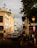 A busy urban street lined with colorful colonial buildings on both sides. The buildings showcase intricate architectural details and hues of yellow, blue, and white. Cars are lined up along the cobblestone street, and people are walking on the sidewalks. A sign for traffic, 'Transito' and 'PARE', is visible along with orange traffic cones. Overhead, there are lamps and electrical wires, and trees add greenery to the scene.