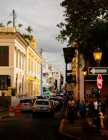A busy urban street lined with colorful colonial buildings on both sides. The buildings showcase intricate architectural details and hues of yellow, blue, and white. Cars are lined up along the cobblestone street, and people are walking on the sidewalks. A sign for traffic, 'Transito' and 'PARE', is visible along with orange traffic cones. Overhead, there are lamps and electrical wires, and trees add greenery to the scene.