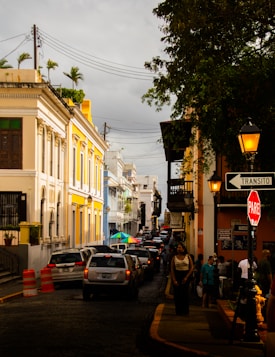 A busy urban street lined with colorful colonial buildings on both sides. The buildings showcase intricate architectural details and hues of yellow, blue, and white. Cars are lined up along the cobblestone street, and people are walking on the sidewalks. A sign for traffic, 'Transito' and 'PARE', is visible along with orange traffic cones. Overhead, there are lamps and electrical wires, and trees add greenery to the scene.