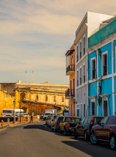 A street scene with colonial-style architecture, featuring colorful buildings including a bright blue one. Several cars are parked along the side of the narrow road, which leads up to a historical fortress with weathered stone walls. People are walking along the street, and the sky is a blend of soft, warm colors.