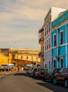 A street scene with colonial-style architecture, featuring colorful buildings including a bright blue one. Several cars are parked along the side of the narrow road, which leads up to a historical fortress with weathered stone walls. People are walking along the street, and the sky is a blend of soft, warm colors.