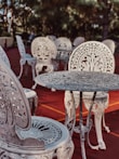 Intricately designed ornate metal chairs and tables are arranged on a reddish outdoor surface. The furniture features elaborate floral patterns and elegant craftsmanship, set against a backdrop of blurred greenery and trees.
