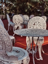Intricately designed ornate metal chairs and tables are arranged on a reddish outdoor surface. The furniture features elaborate floral patterns and elegant craftsmanship, set against a backdrop of blurred greenery and trees.