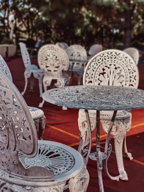 Intricately designed ornate metal chairs and tables are arranged on a reddish outdoor surface. The furniture features elaborate floral patterns and elegant craftsmanship, set against a backdrop of blurred greenery and trees.