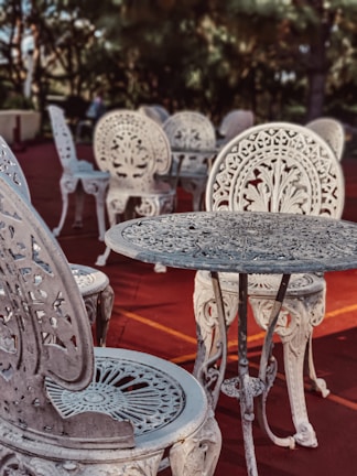 Intricately designed ornate metal chairs and tables are arranged on a reddish outdoor surface. The furniture features elaborate floral patterns and elegant craftsmanship, set against a backdrop of blurred greenery and trees.