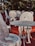 Intricately designed ornate metal chairs and tables are arranged on a reddish outdoor surface. The furniture features elaborate floral patterns and elegant craftsmanship, set against a backdrop of blurred greenery and trees.