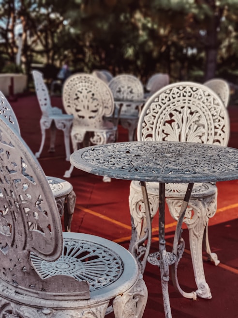 Intricately designed ornate metal chairs and tables are arranged on a reddish outdoor surface. The furniture features elaborate floral patterns and elegant craftsmanship, set against a backdrop of blurred greenery and trees.