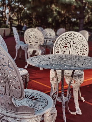 Intricately designed ornate metal chairs and tables are arranged on a reddish outdoor surface. The furniture features elaborate floral patterns and elegant craftsmanship, set against a backdrop of blurred greenery and trees.
