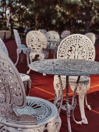 Intricately designed ornate metal chairs and tables are arranged on a reddish outdoor surface. The furniture features elaborate floral patterns and elegant craftsmanship, set against a backdrop of blurred greenery and trees.