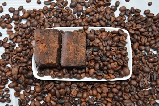 A white rectangular dish is filled with coffee beans and two blocks of ground coffee. Coffee beans are scattered around the dish on a light-colored surface.