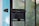 A rusted black latch is attached to a metal mesh screen door. The metal door frames are black, and a portion of a light blue painted wooden door frame is visible on the left. The screen allows a slightly blurred view of greenery outside, suggesting an outdoor environment beyond the door.