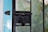 A rusted black latch is attached to a metal mesh screen door. The metal door frames are black, and a portion of a light blue painted wooden door frame is visible on the left. The screen allows a slightly blurred view of greenery outside, suggesting an outdoor environment beyond the door.