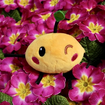 A cute, round, plush toy with one eye winking sits among vibrant pink flowers with yellow centers. The toy has a cheerful expression, with a small green leaf perched on top and rosy cheeks.