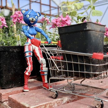 A toy bear with a blue body and red and white striped clothing is positioned on a wooden surface. It is pushing a small shopping cart filled with a black flower pot. In the background, there are several potted pink flowers and greenery, suggesting a garden or nursery setting.