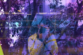 The image captures a reflection of a person taking a photo, with a backdrop of a surreal and vibrant urban scene. The view includes trees, a building with windows, and a mix of natural and artificial lighting blending to create a colorful, abstract composition.