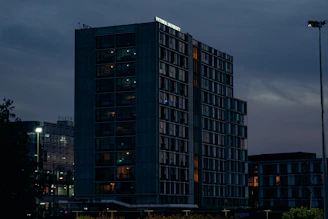 A dramatic dusk view of a contemporary building with glowing interiors and reflective glass, framed by subtle city lights.