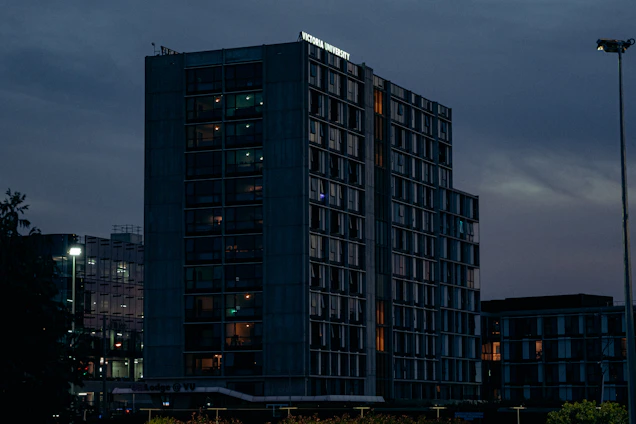 A sleek industrial building at dusk with warm interior lights glowing through large windows, set against a dark gray sky.