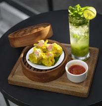 A wooden board holds a bamboo steamer basket containing a plate of yellow dumplings. Next to the basket, there is a small white bowl filled with a red sauce. A refreshing drink in a tall glass garnished with mint leaves and a lime slice is positioned nearby. The setting suggests a casual dining experience.