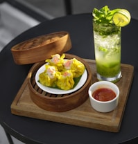A wooden board holds a bamboo steamer basket containing a plate of yellow dumplings. Next to the basket, there is a small white bowl filled with a red sauce. A refreshing drink in a tall glass garnished with mint leaves and a lime slice is positioned nearby. The setting suggests a casual dining experience.