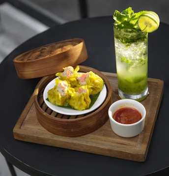 A wooden board holds a bamboo steamer basket containing a plate of yellow dumplings. Next to the basket, there is a small white bowl filled with a red sauce. A refreshing drink in a tall glass garnished with mint leaves and a lime slice is positioned nearby. The setting suggests a casual dining experience.