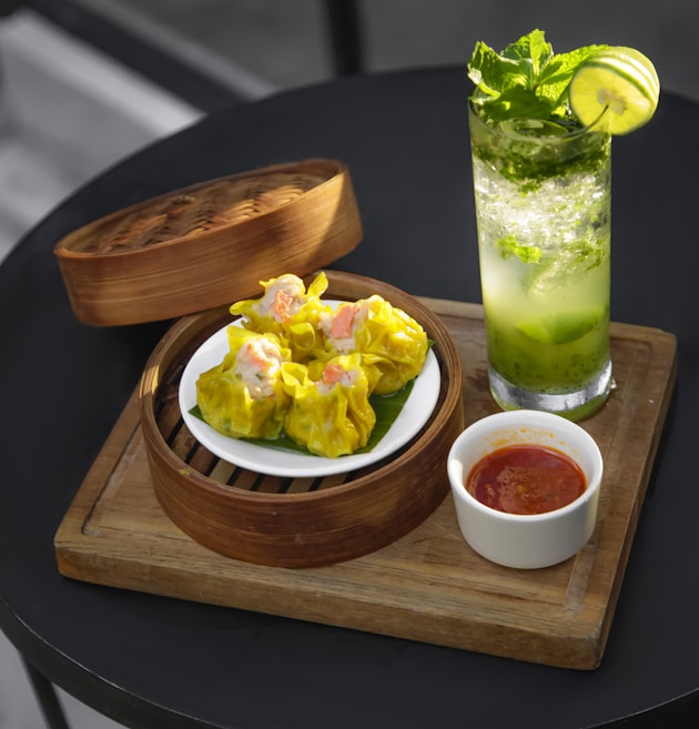 A wooden board holds a bamboo steamer basket containing a plate of yellow dumplings. Next to the basket, there is a small white bowl filled with a red sauce. A refreshing drink in a tall glass garnished with mint leaves and a lime slice is positioned nearby. The setting suggests a casual dining experience.