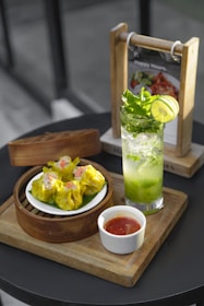 A serving of vibrant yellow dim sum arranged in a bamboo steamer with a side of red dipping sauce in a small white cup. Next to it, a refreshing green mint and lime drink sits in a clear glass. The setup is on a wooden tray, with a menu holder visible in the background.