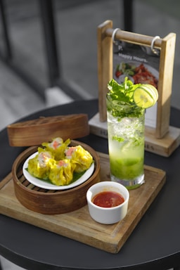 A serving of vibrant yellow dim sum arranged in a bamboo steamer with a side of red dipping sauce in a small white cup. Next to it, a refreshing green mint and lime drink sits in a clear glass. The setup is on a wooden tray, with a menu holder visible in the background.