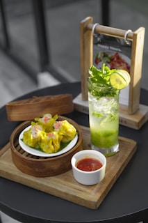 A serving of vibrant yellow dim sum arranged in a bamboo steamer with a side of red dipping sauce in a small white cup. Next to it, a refreshing green mint and lime drink sits in a clear glass. The setup is on a wooden tray, with a menu holder visible in the background.