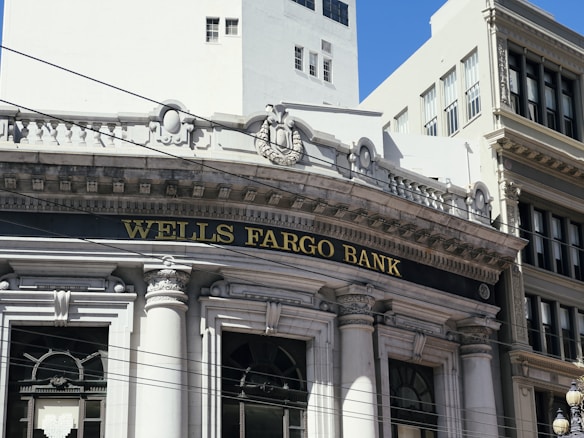 The image depicts the facade of a historic building with classical architectural elements, including ornate carvings and columns. The prominently displayed name 'Wells Fargo Bank' is set against a black background above large arched windows.