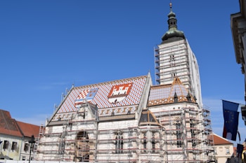 A historical building with a vibrant, decorative roof displaying colorful tiles that form patterns and coats of arms. The structure is under renovation, evident from the scaffolding surrounding it. The sky is clear and blue, adding to the visual appeal.
