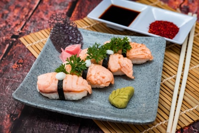 A plate of sushi with salmon nigiri topped with a white sauce and garnished with fresh parsley. Accompanied by wasabi, pickled ginger, and a decorative leaf on a textured gray plate. Dipping sauces are arranged in a small rectangular dish beside the sushi, all placed on bamboo mats.