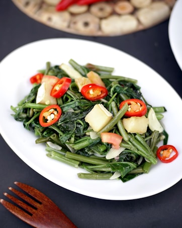 A plate of stir-fried vegetables featuring green leafy stems, red chili slices, and chunks of potato. The dish is presented on a white oval plate with a dark background, alongside a wooden fork. The vibrant colors of the vegetables stand out, creating an appetizing appearance.