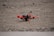 A small quadcopter drone with red propellers is positioned on a gravel-covered surface. It features a compact design with visible electronic and mechanical components mounted on a yellow and black battery pack.