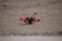 A small quadcopter drone with red propellers is positioned on a gravel-covered surface. It features a compact design with visible electronic and mechanical components mounted on a yellow and black battery pack.
