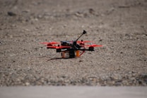A small quadcopter drone with red propellers is positioned on a gravel-covered surface. It features a compact design with visible electronic and mechanical components mounted on a yellow and black battery pack.