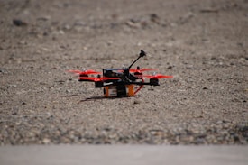 A small quadcopter drone with red propellers is positioned on a gravel-covered surface. It features a compact design with visible electronic and mechanical components mounted on a yellow and black battery pack.