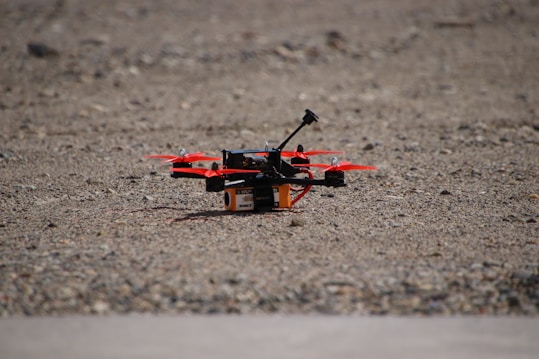 A small quadcopter drone with red propellers is positioned on a gravel-covered surface. It features a compact design with visible electronic and mechanical components mounted on a yellow and black battery pack.