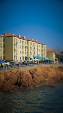 A row of light-colored apartment buildings with red-tiled roofs is situated alongside a rocky waterfront. Several small shops or cafes with blue awnings line the street below these buildings. People and vehicles can be seen along the promenade, and the scene is under a clear blue sky.