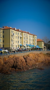 A row of light-colored apartment buildings with red-tiled roofs is situated alongside a rocky waterfront. Several small shops or cafes with blue awnings line the street below these buildings. People and vehicles can be seen along the promenade, and the scene is under a clear blue sky.