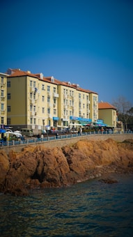 A row of light-colored apartment buildings with red-tiled roofs is situated alongside a rocky waterfront. Several small shops or cafes with blue awnings line the street below these buildings. People and vehicles can be seen along the promenade, and the scene is under a clear blue sky.