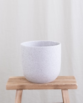 A minimalist ceramic pot with a speckled texture is placed on a simple wooden stool. The background is a plain, neutral-toned fabric.