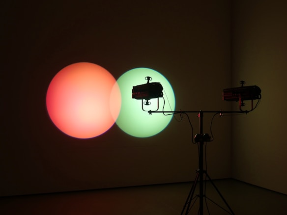Two spotlights create overlapping circles of light on a wall, one red and one green, mounted on a metal tripod stand.