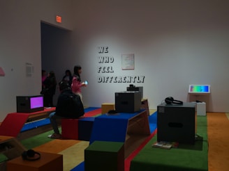 The image depicts an interior space with several people interacting with technological displays. The wall features the phrase 'We Who Feel Differently,' and various benches are covered in colorful fabric. Headphones are placed on benches near monitors.