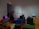 The image depicts an interior space with several people interacting with technological displays. The wall features the phrase 'We Who Feel Differently,' and various benches are covered in colorful fabric. Headphones are placed on benches near monitors.