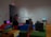 The image depicts an interior space with several people interacting with technological displays. The wall features the phrase 'We Who Feel Differently,' and various benches are covered in colorful fabric. Headphones are placed on benches near monitors.