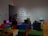 The image depicts an interior space with several people interacting with technological displays. The wall features the phrase 'We Who Feel Differently,' and various benches are covered in colorful fabric. Headphones are placed on benches near monitors.
